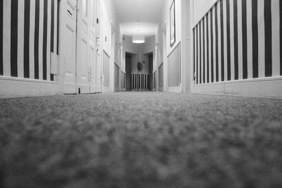 Low-angle view of a corridor with textured carpet flooring, flanked by white wooden paneling and doors on the left side, and vertical striped wallpaper on the right side. The corridor is illuminated b
