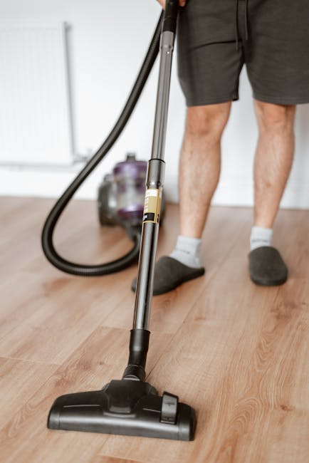 A woman using a yellow vacuum cleaner to deep clean a patterned beige and green rug in a living room with wooden flooring and a red leather sofa, demonstrating surface cleaning and maintenance. The ro